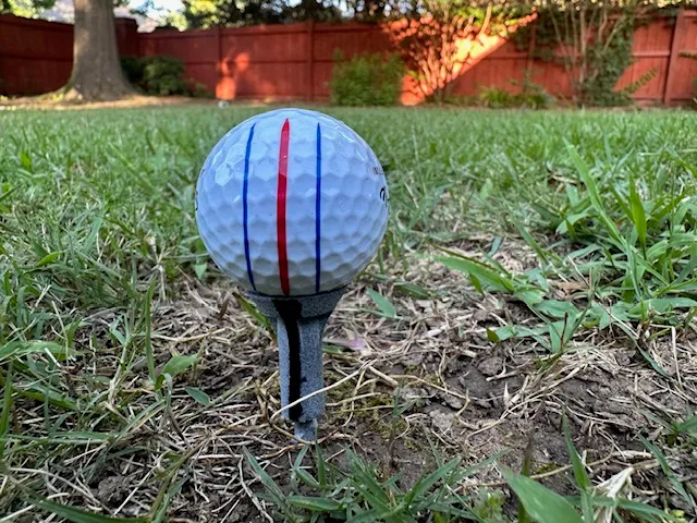 A lined golf ball resting on the red Noisiv Universal adjustable tee, set in fairway grass.